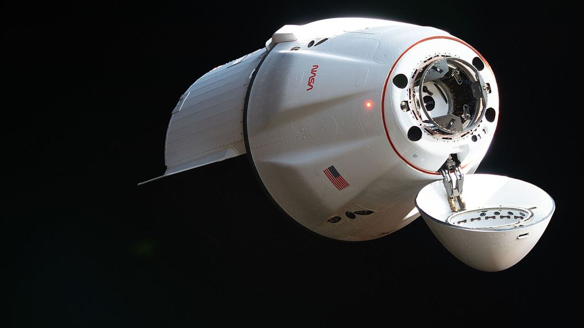 A white SpaceX cargo Dragon with an American flag and its nose cone open glides against black space