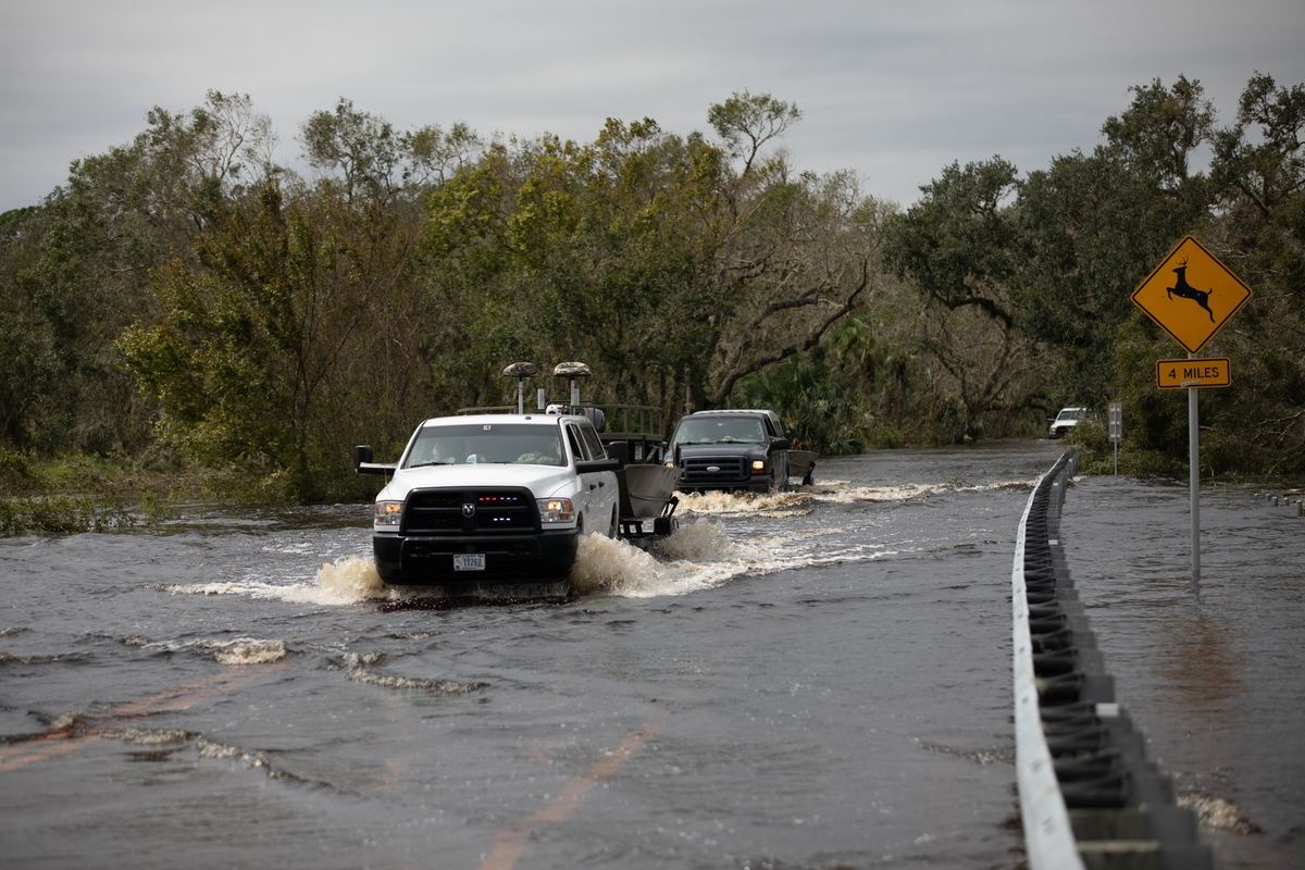 Pickup trucks towing rescue boats drive through a flooded Florida road lined with green trees