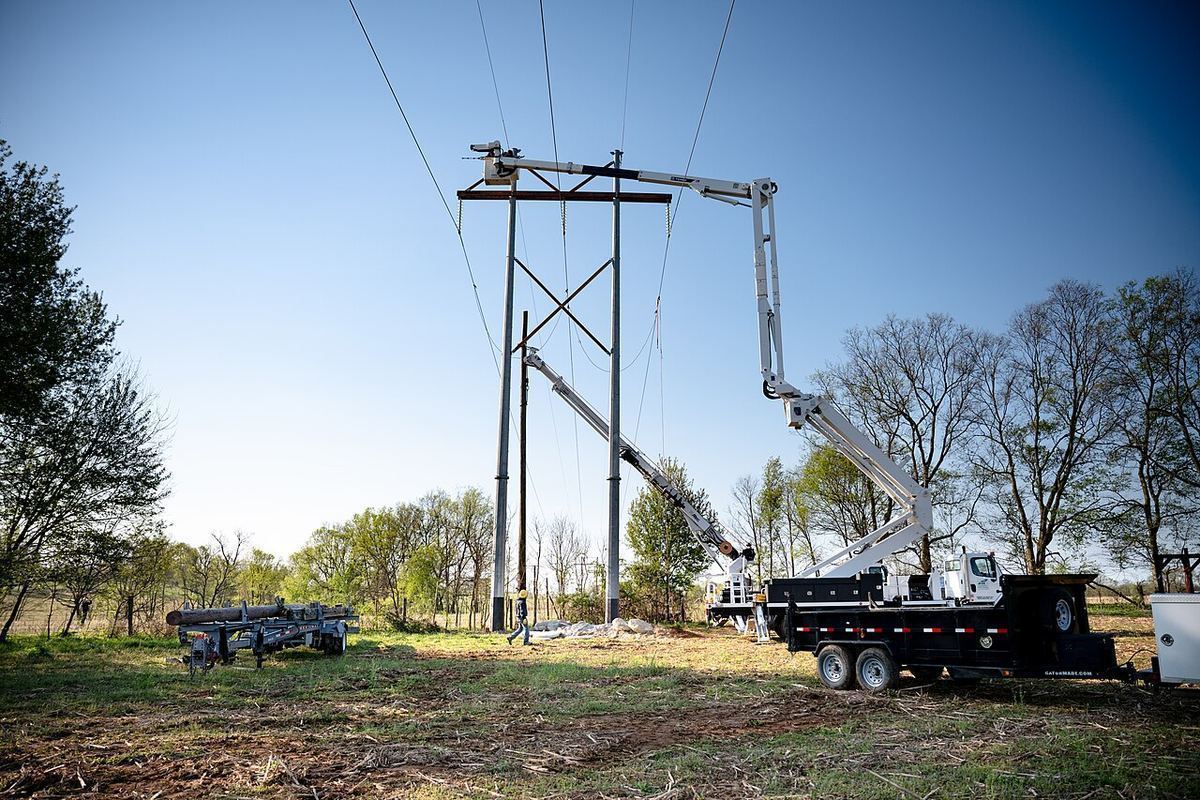 Bucket trucks raising a new power line across a green rural field under a clear spring sky