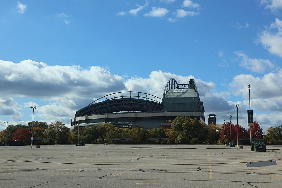 American Family Field's arched roof under a blue autumn sky, seen across an empty parking lot lined with red and gold trees