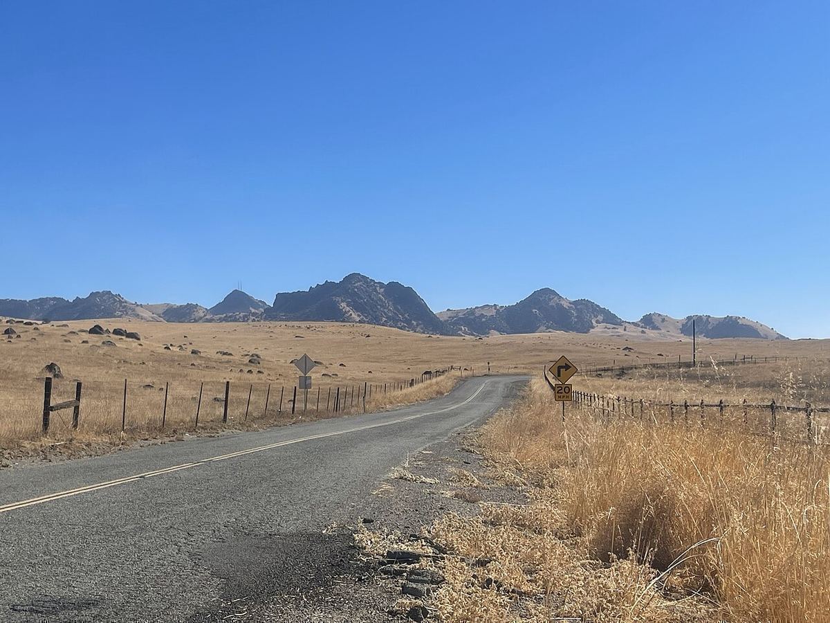 A quiet two-lane country road runs through golden autumn grassland toward the rugged Sutter Buttes under a clear blue sky